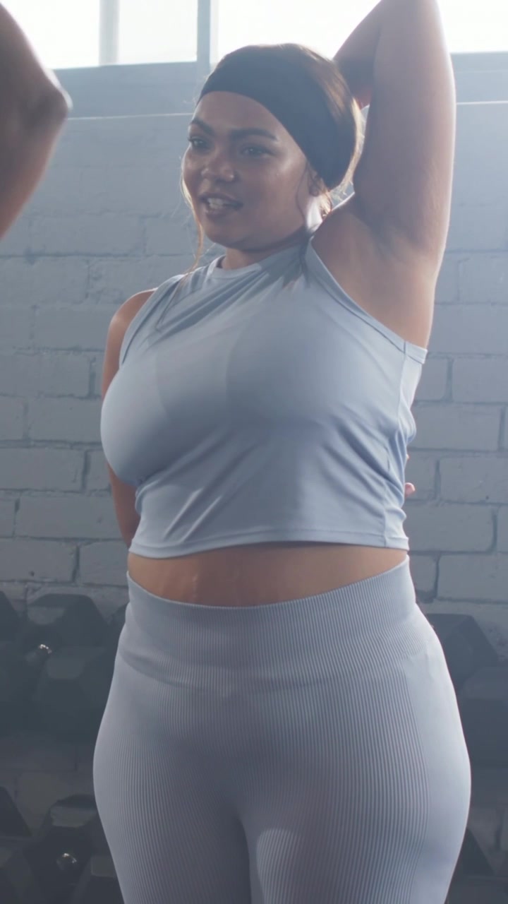 Video verticale: Un istruttore che guida una donna mentre allunga i tricipiti in palestra, vicino a una rastrelliera di manubri, con un top blu.