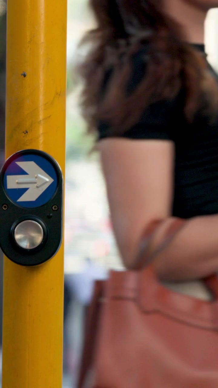 Video verticale: Donna in attesa con top nero e borsa color cuoio al marciapiede, vicino a un palo giallo, guarda il semaforo cambiare per attraversare.