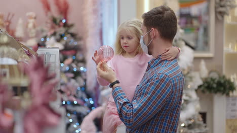 a father in a medical mask on his face with his daughter choose jewelry and toys on christmas eve to decorate the house and the christmas tree