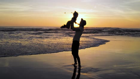 Una-Madre-Disfruta-De-Las-Vacaciones-Con-Su-Hijo-Durante-La-Puesta-De-Sol-En-La-Playa-Mientras-Lanza-A-Su-Hijo-Al-Aire-Con-Alegría