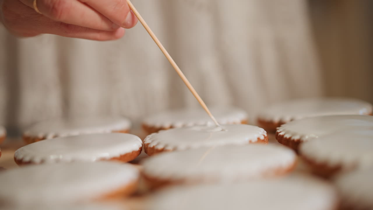 Macro di una mano caucasica che applica precise pennellate di glassa alla zucca su biscotti rotondi, artigiano pasticcere concentrato che crea motivi uniformi con uno stuzzicadenti, luce da studio in cucina, tecnica meditativa che cattura la texture.