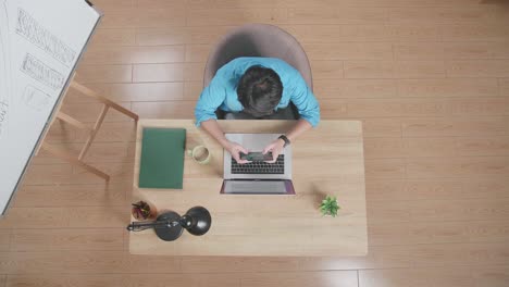 top view of a man playing game on smartphone while sitting at the wooden desk besides a whiteboard typing on a laptop at home