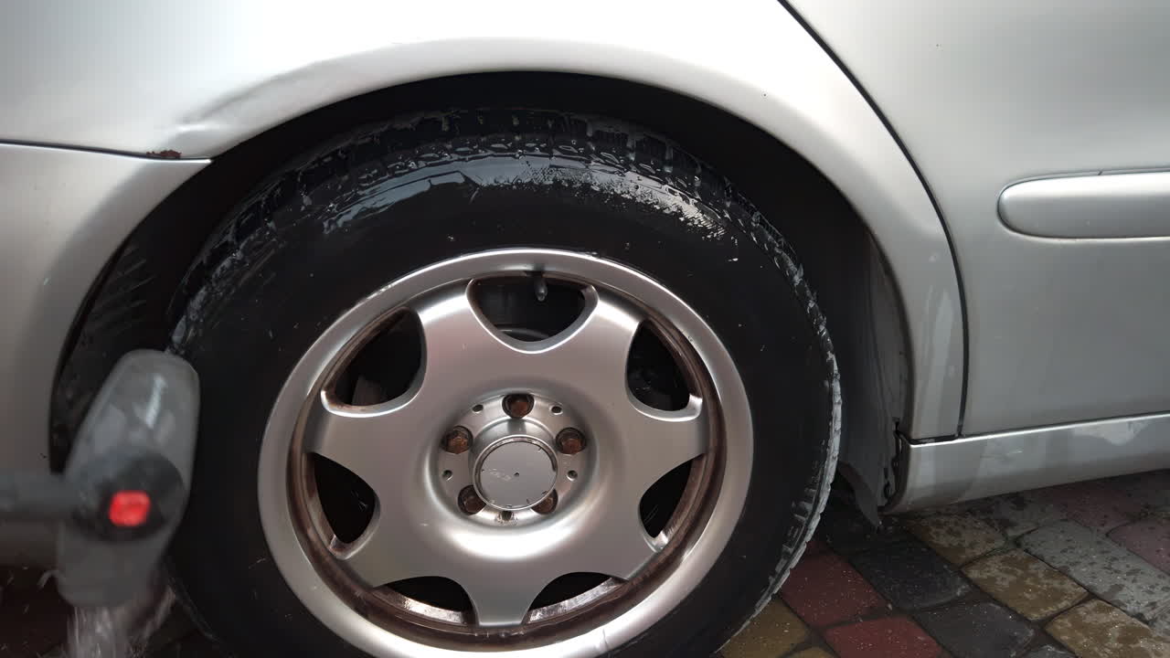 Cleaning the car tires and wheels with water and soap in a residential driveway during a rainy day. The focus is on removing dirt and grime for a refreshed look