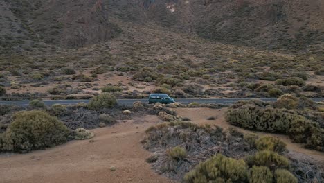 Aerial-Following-Shot-as-a-Cyan-Car-Drives-Down-Highway-Filled-Covered-by-Shrubs-and-Brush,-Tenerife-Spain