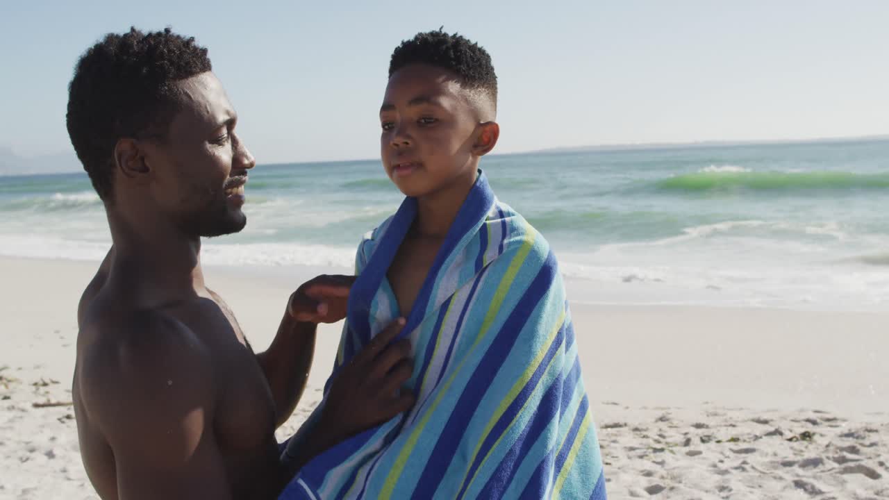Padre afroamericano sorridente che asciuga il figlio su una spiaggia assolata.
