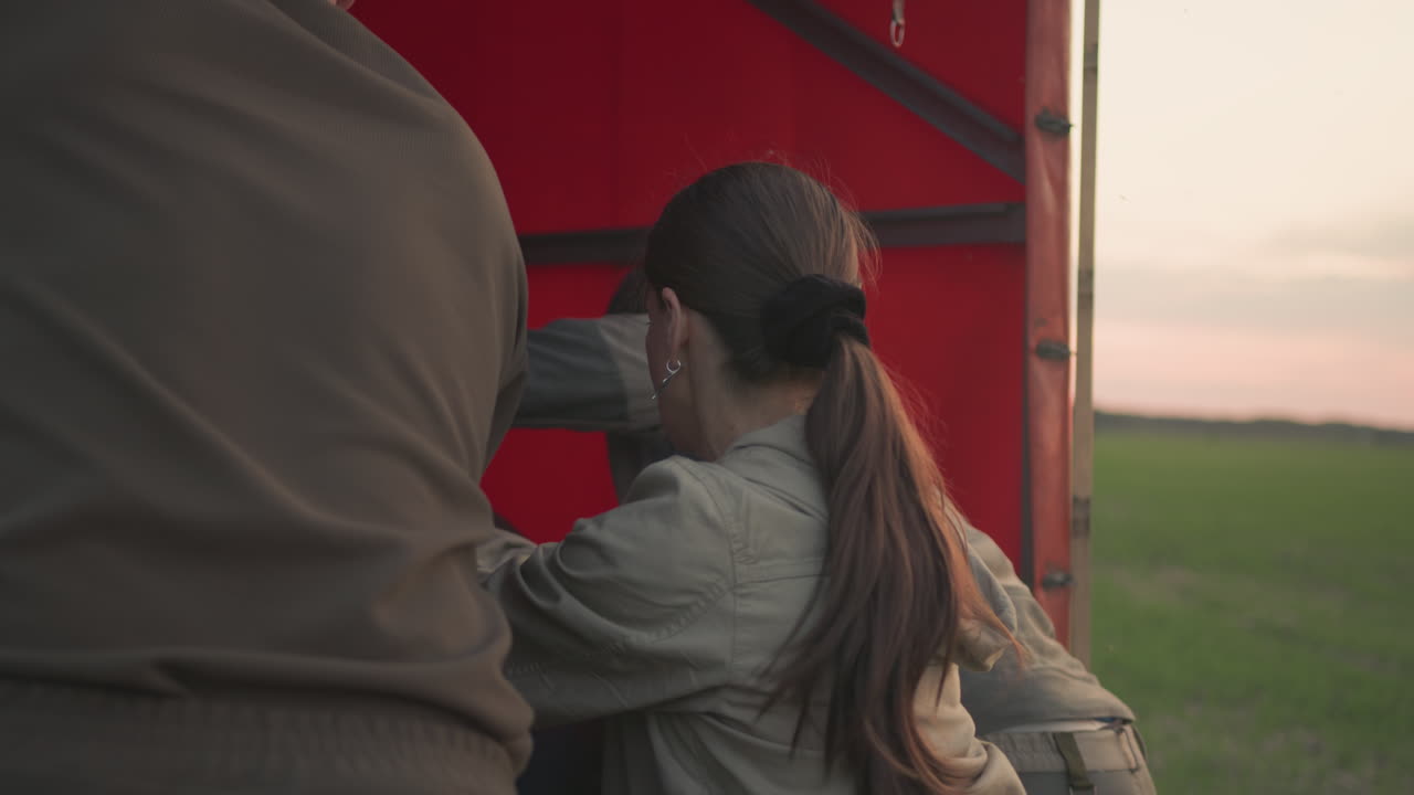 close up of three people pushing oversized metal burner coil assembly into red trailer bed at dusk on wide farm field, highlighting coordinated effort, hand gripping motion