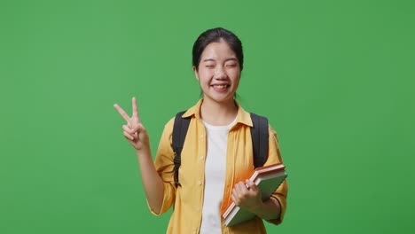 asian woman student with a backpack and some books smiling and showing peace gesture while standing in the green screen background studio