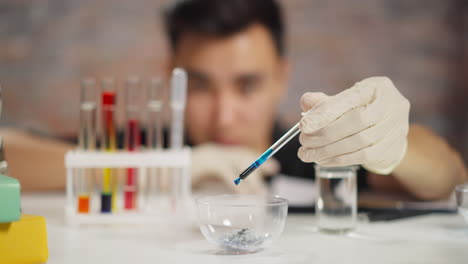 Chemist-with-gloves-drops-blue-reagent-in-bowl-with-crystals