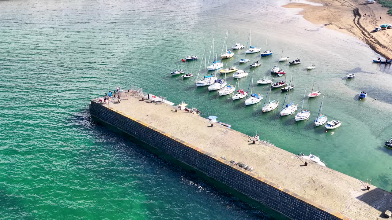 Drone camera glides over marina with sailboats, stone pier, and clear summer coastal waters