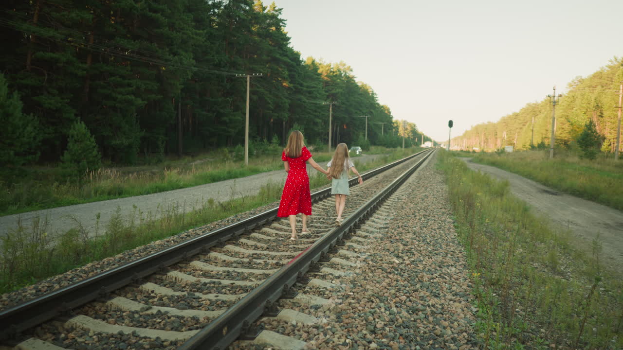 mother in red dress holds little girl hand for balance as they walk along rail track through rural area with forest trees, gravel path, parked car, and utility poles in background on sunny day
