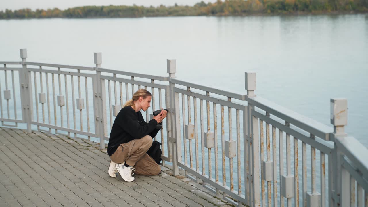 Ung kvinde hukker sig ned på en mole og fanger kystlinjen med telefon, iført sort jakke, khakibukser og hvide sneakers, siddende på et trædæk bag et metalgelænder, rolig overskyet aften, eftertænksom