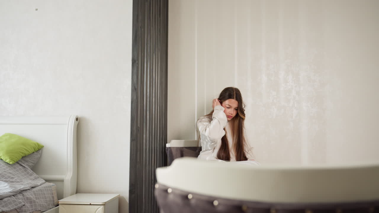 Student walks past bed with divider, moves to seat, sets nail polish on table, adjusts long hair, picks polish again, studies bottle in quiet room, soft daylight, tidy bedding with green pillows