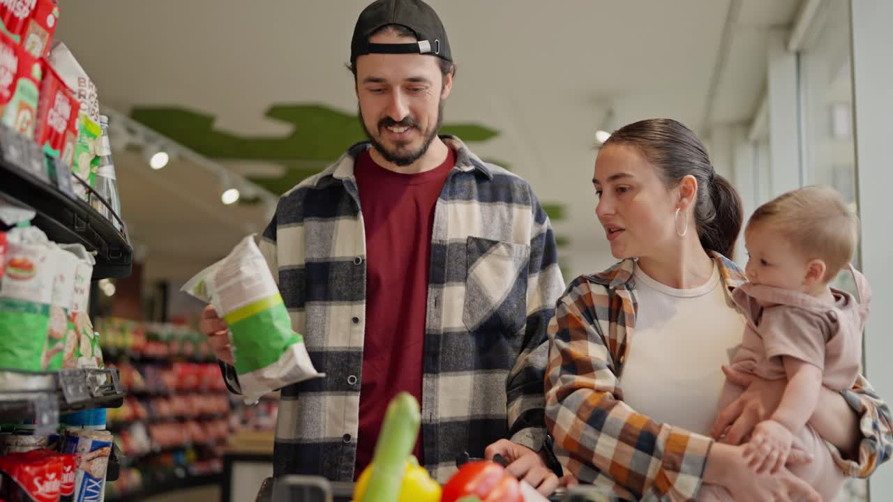 A confident brunette woman in a checkered shirt advises what to choose for her husband during family shopping with a small child in the supermarket