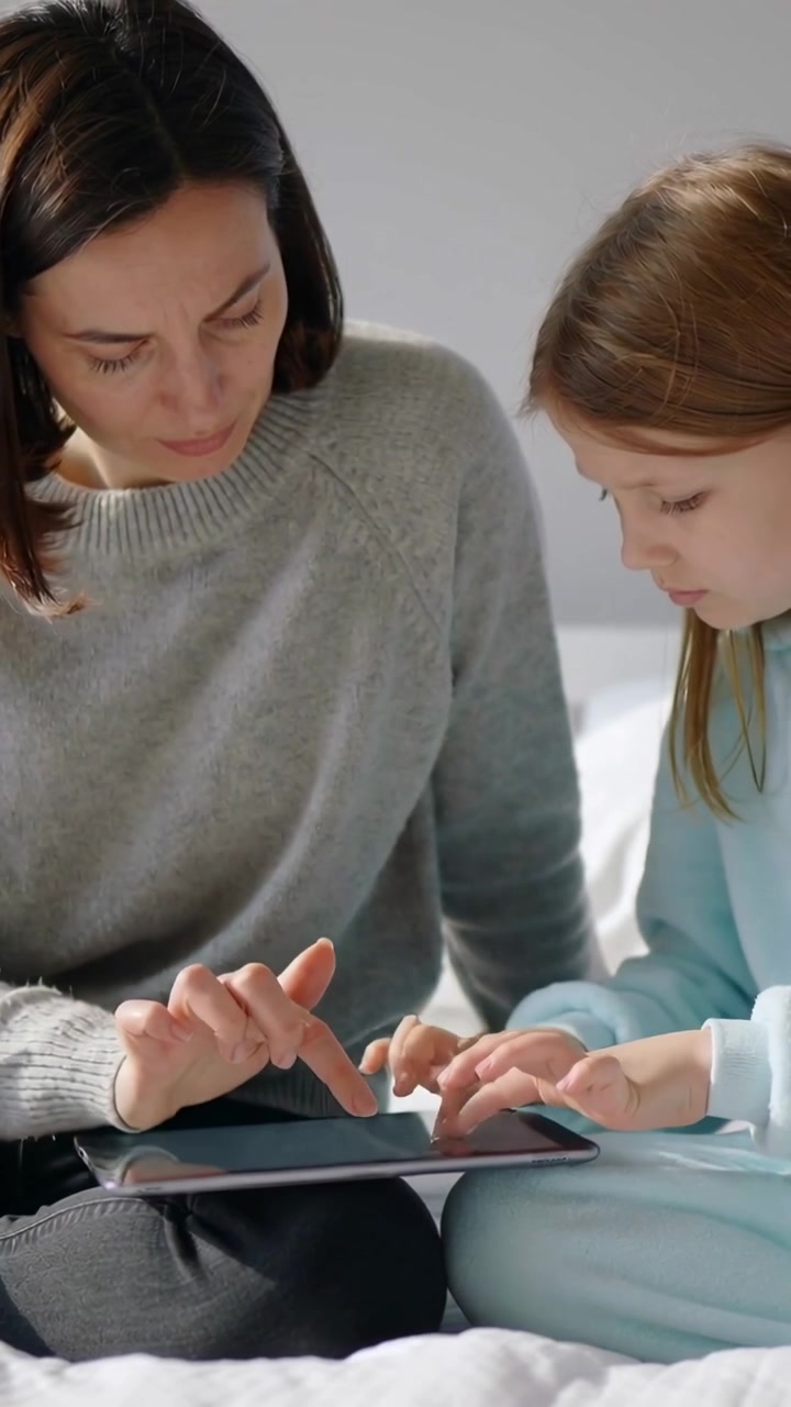 Video verticale: Mamma in maglione grigio che si china insegnando al bambino in abiti blu a scorrere un tablet sul letto