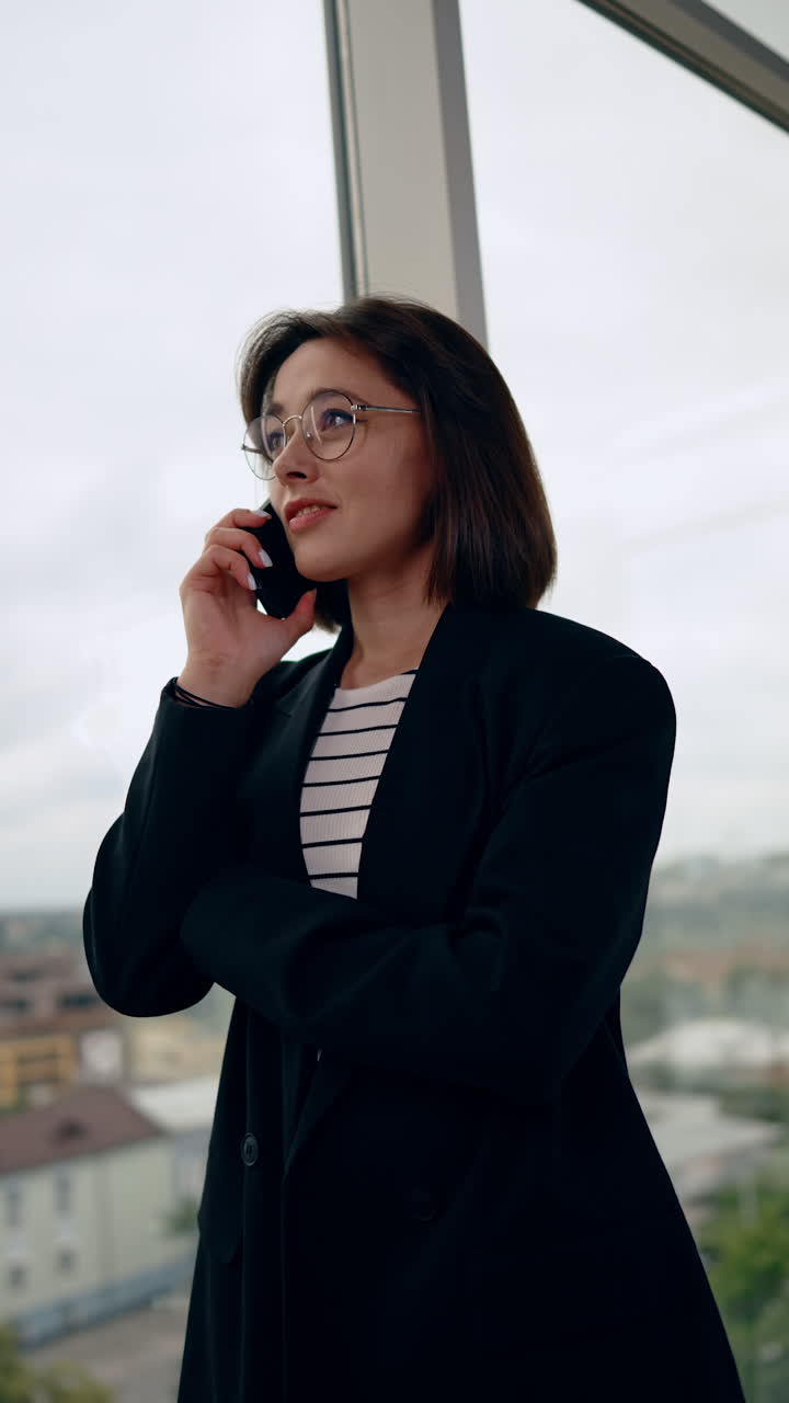 Young Caucasian brunette woman in black jacket stands at the window. Lady in glasses talks on the phone looking at window. Low angle view. Vertical video.