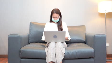 dressed in loose-fitting clothes and wearing a surgical mask, a young woman works from home on her laptop