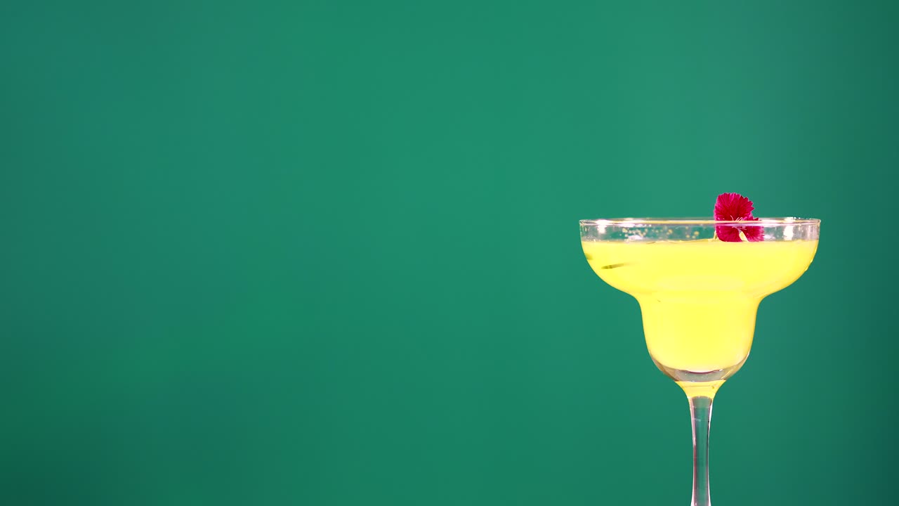 A vibrant yellow cocktail with a cherry garnish against a green screen backdrop, showcasing a static, well-lit presentation