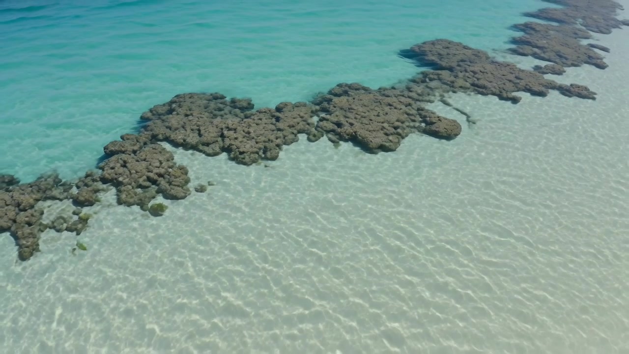 Svævende drone filmer sollys, der skifter og afslører spredte revpletter over bleg sandbund.