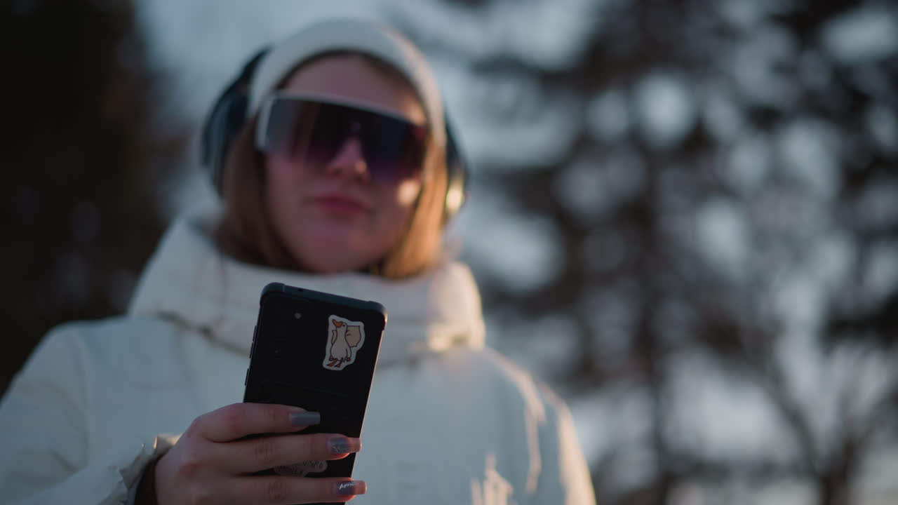 Close up of woman wearing puffer coat and tinted goggles pressing smartphone with long manicured nails under soft winter light against blurred forest backdrop conveying modern lifestyle vibe