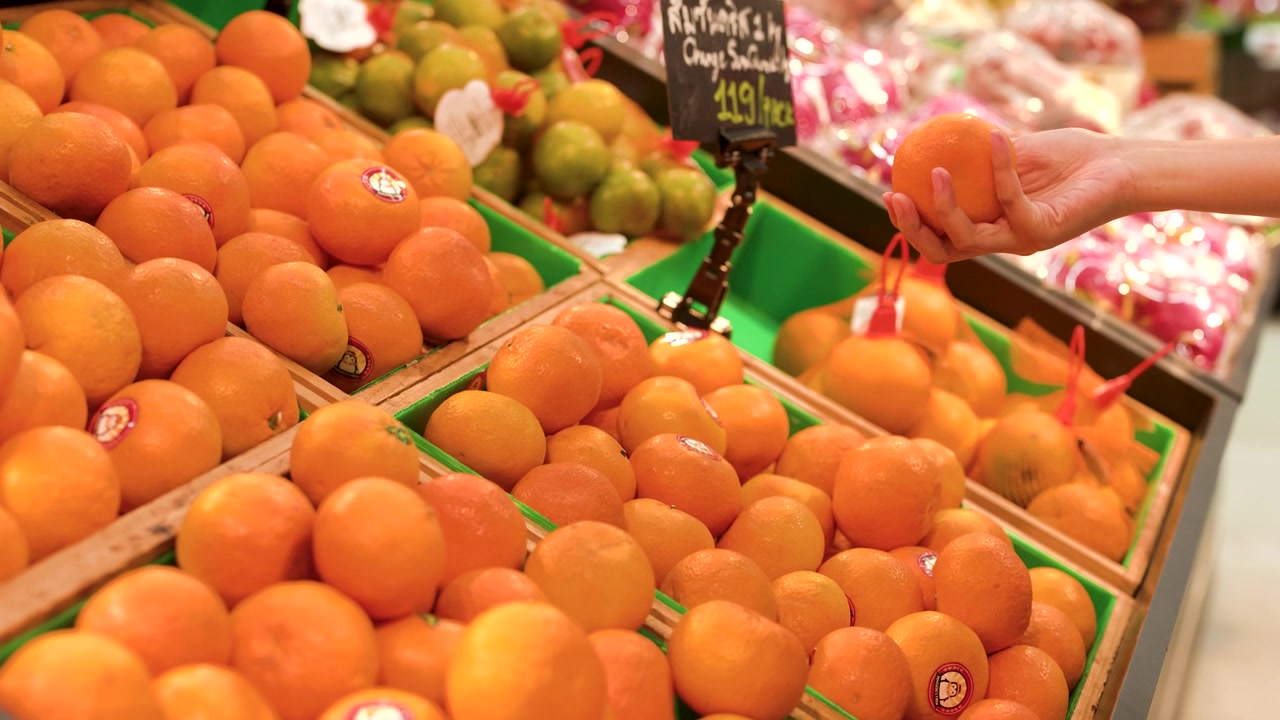 Close-up of a hand picking a ripe orange from a grocery store fruit bin.