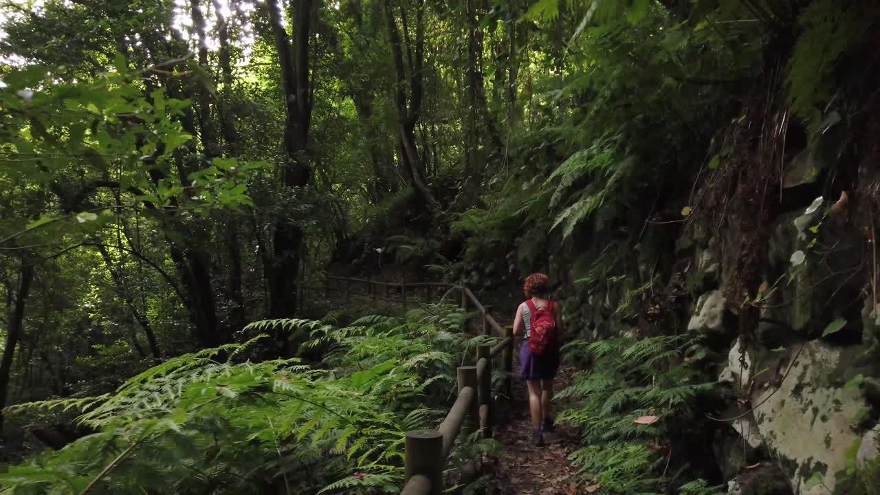 A shot following a female hiker walking on a narrow path with a fence