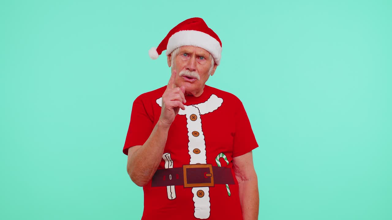 Senior old man in christmas tshirt presses index finger to lips makes silence gesture sign secret