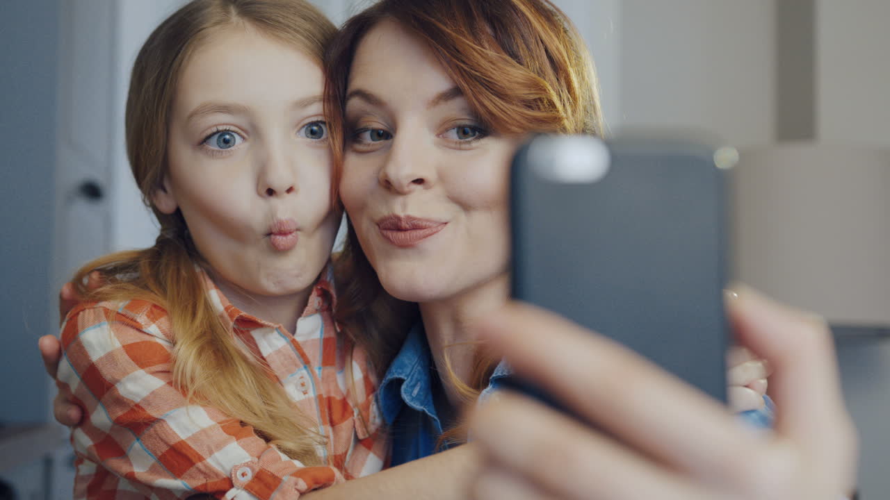 Close up of the smiling mother hugging young daughter and making funny selfies on the smartphone while grimacing. Portrait. Inside