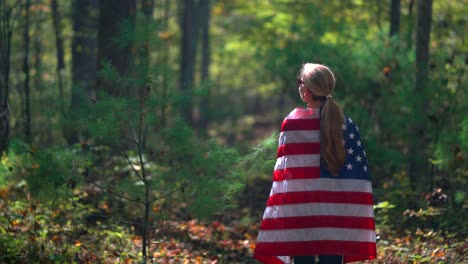 blonde woman with flag wrapped around her walking through a sun lit forest