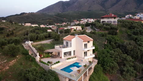 aerial pull away from luxury greek villa with pool revealing traditional greek village in background - car on drive