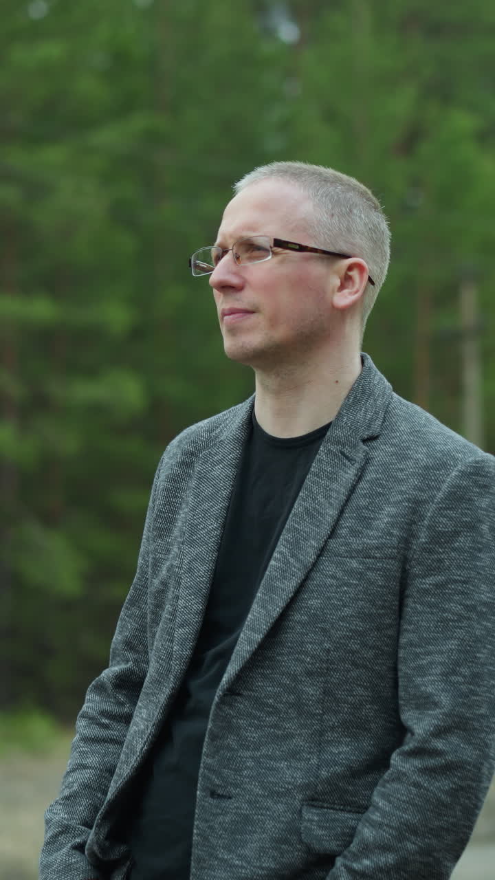 Close-up view of a man in glasses and a gray jacket standing on a railway track, removed his glasses with one hand while looking thoughtfully into the distance, with a blurred background