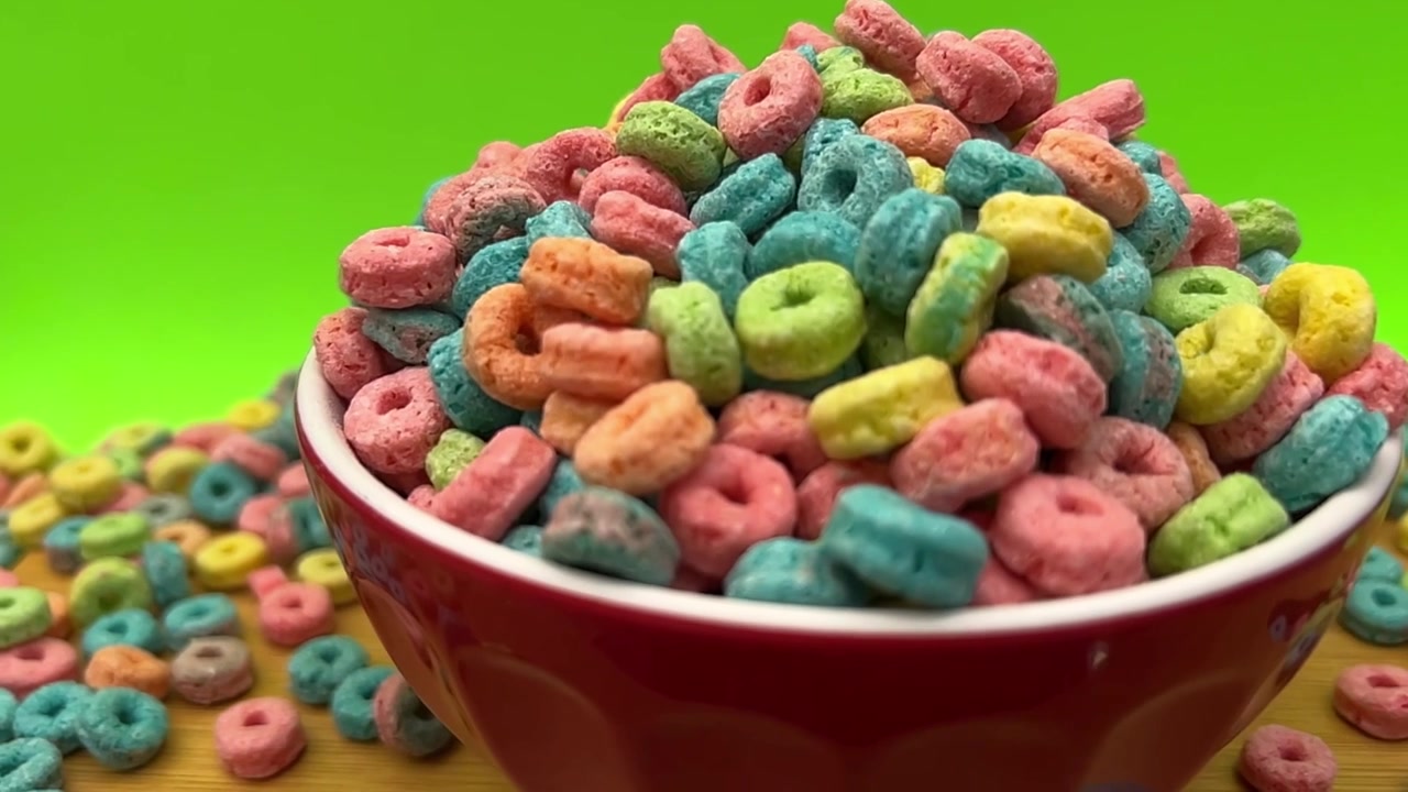 Anneaux de céréales colorés empilés dans un bol rouge sur un fond vert vif, montrant des morceaux de céréales de petit-déjeuner assortis en rose, bleu, jaune, orange et vert