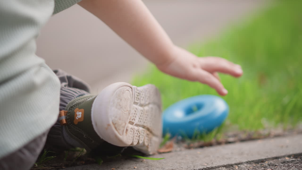 Un bambino trova un giocattolo. Un bambino piccolo esplora l'ambiente esterno. Un bambino esamina un anello blu sul bordo di un sentiero erboso. Un giovane esploratore allunga la mano verso un giocattolo blu su un tranquillo bordo erboso durante l'estate.