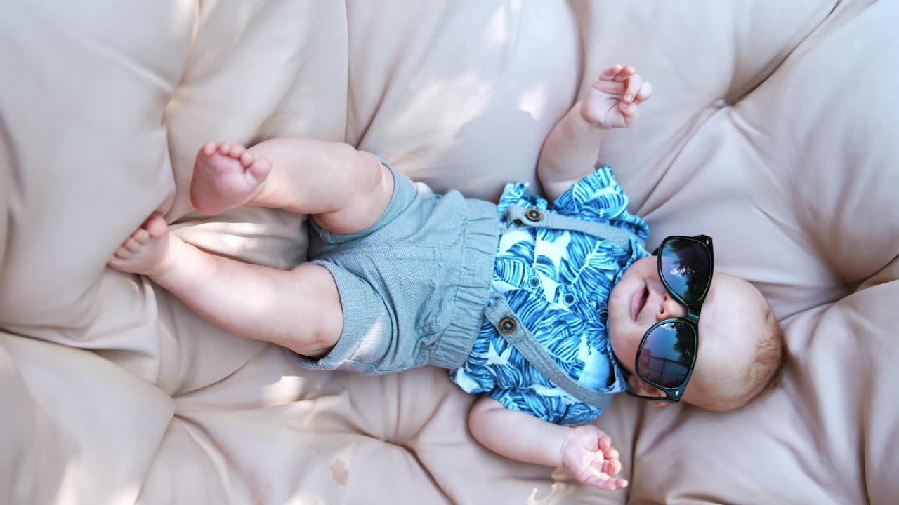 Cute baby boy wearing grey shorts with suspenders and blue shirt lies on the soft sofa. Infant is wearing adult sunglasses. Top view. Vertical screen.