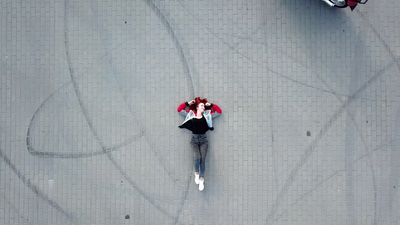 red-haired woman in sportswear is laying in the middle of pavement and surrounded by two motorcycles that are riding around her. Camera motion up