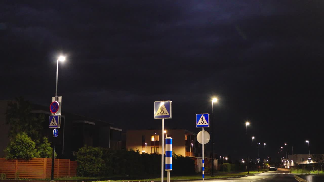 Low angle wide shot video of a dark night thunder sky with lighting strikes visible in a quiet and modern rural neighbourhood where apartment buildings and street light illuminated streets are visible