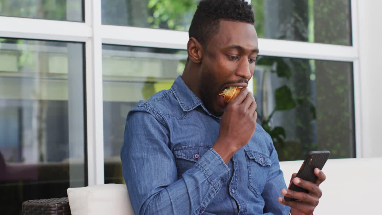 Uomo d'affari afroamericano che usa lo smartphone e mangia un croissant in un bar