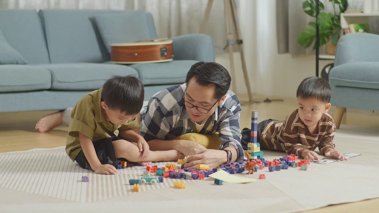Full Body Of Asian Father And Sons Playing The Construction Set Colorful Plastic Toy Brick At Home. The Kids Assemble Plastic Building Blocks Sitting On A Mat, A Father Lying On The Floor Talking To His Sons