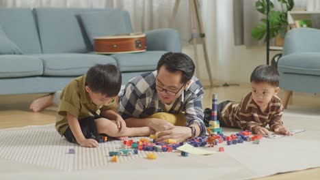 full body of asian father and sons playing the construction set colorful plastic toy brick at home. the kids assemble plastic building blocks sitting on a mat, a father lying on the floor talking to his sons