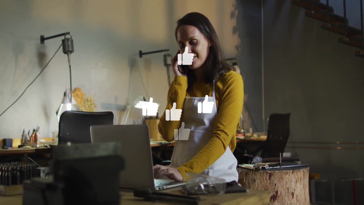 woman talking on smartphone while typing on laptop in workshop, showing animated tech chart overlay