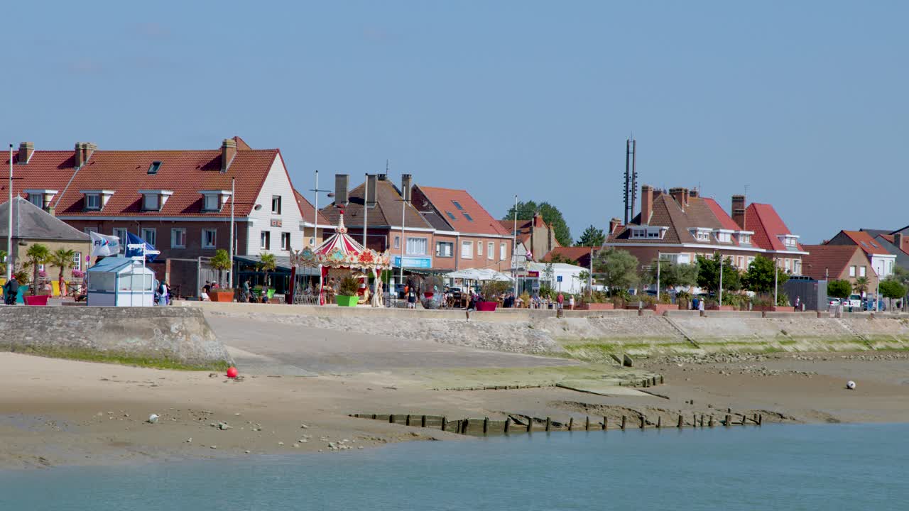 Wide shot slowly pans across seaside town, sandy beach, and calm water under clear daylight