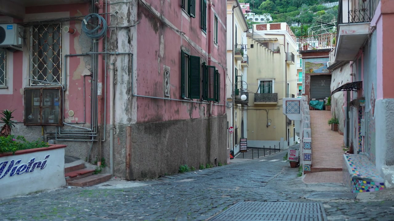 Steep cobblestone alley passes faded pink apartments and tiled stairway in a quiet Vietri Italy side street.