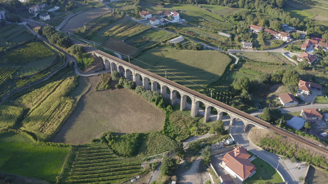 Ripresa aerea orbitale di un vecchio ponte ferroviario ad arco che proietta un'ombra su un campo di mais al tramonto - Ponte Seca, Durrães, Barcelos.