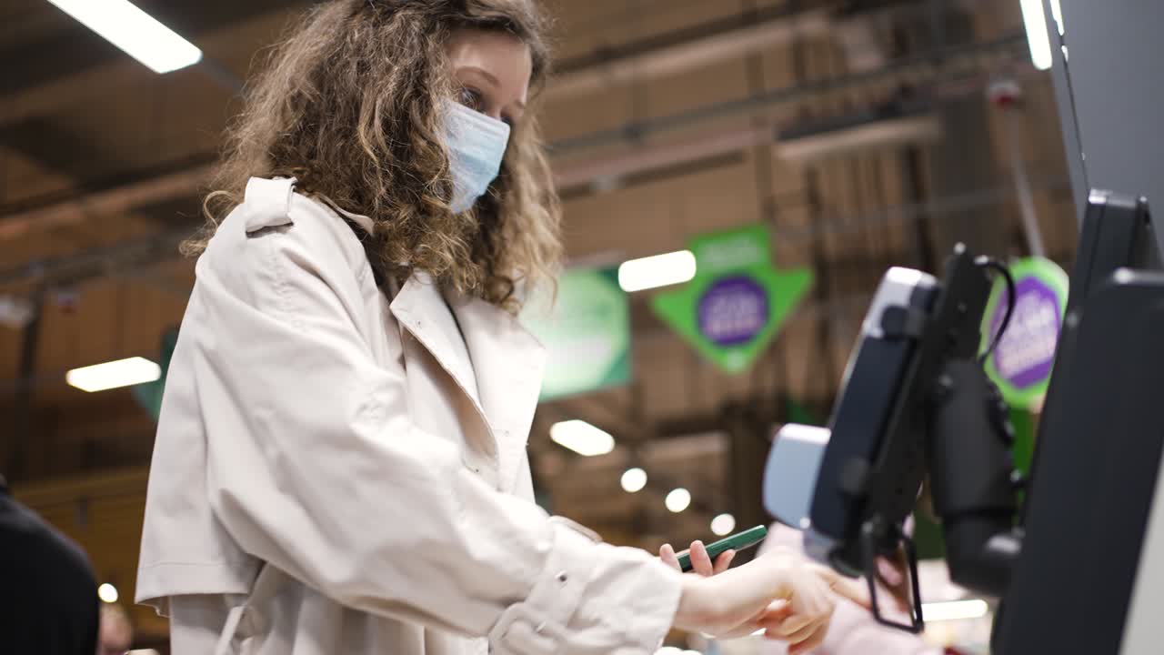 una mujer con una máscara médica hace compras en la caja de autoservicio en un supermercado usando un teléfono móvil