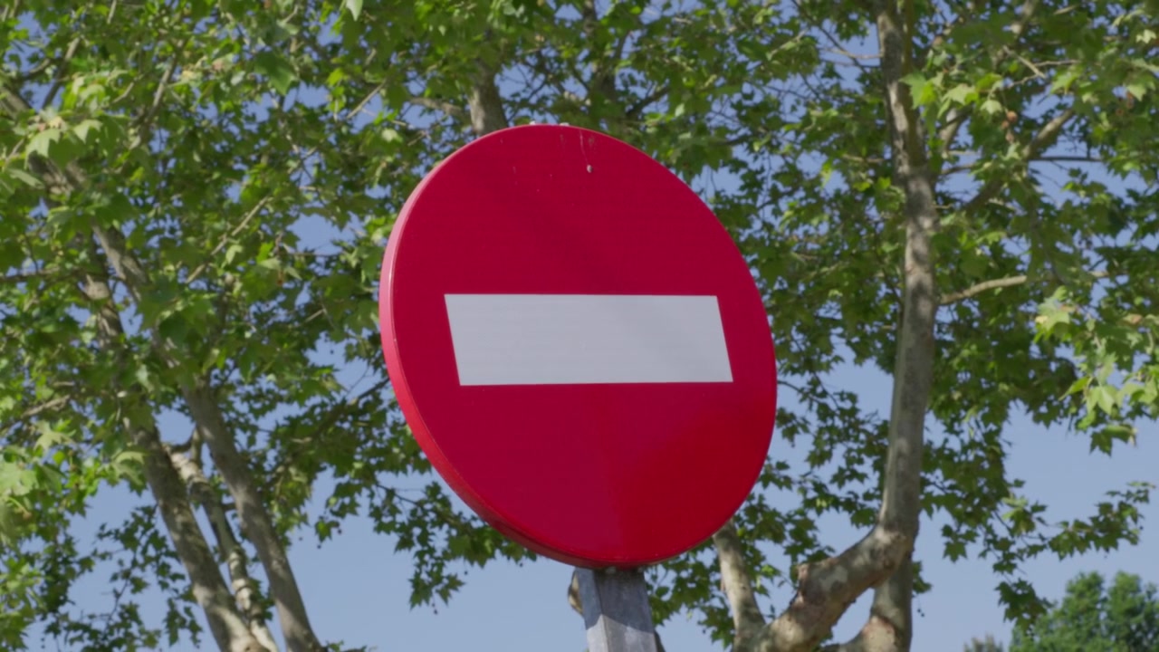 Red no entry traffic sign stands out against leafy green trees and clear blue sky in steady close-up shot, featuring vibrant colors and sharp focus.