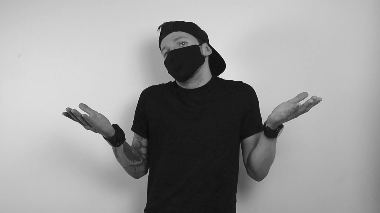 Grayscale footage of a tattooed man wearing a black health protection covid mask and lifting up his arm in confusion like he is questioning something in slow motion. Don't know movement.