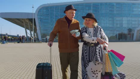 Senior-rich-pensioner-tourists-walking-with-shopping-bags-from-vacation-holding-dollar-money-cash