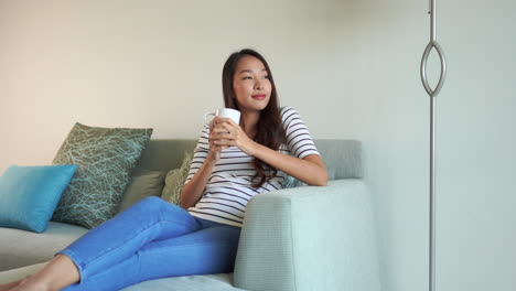 A-young,-pretty,-Asian-woman-dressed-casually-in-jeans-and-a-striped-blouse-reclines-on-a-comfortable-grey-couch-as-she-drinks-a-hot-cup-of-coffee
