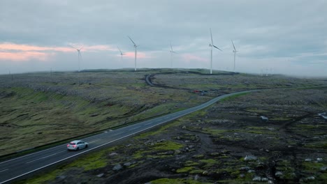 un coche conduce en una carretera panorámica a través de un vasto parque eólico bajo un cielo nublado, evocando la calma