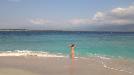 Pretty-young-woman-standing-at-tropical-beach-and-raising-arms-up