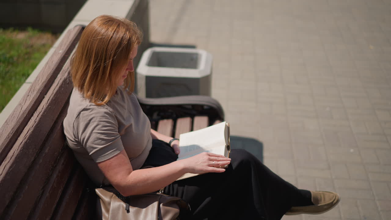 Top view of student sitting on wooden bench reading open book outdoors under bright sunlight, flipping page with calm focus surrounded by warm daylight